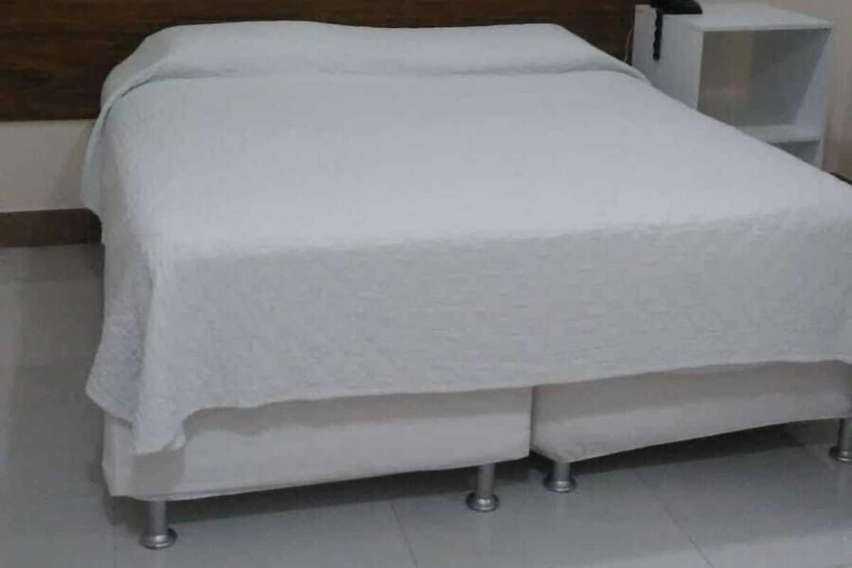 Una cama bien tendida en una habitación de hotel, con cabecera de madera, mesita de noche blanca y un cubo de basura blanco.