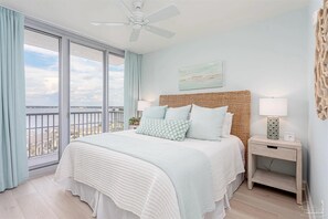 Ein Schlafzimmer mit großem Fenster, Deckenventilator, Bett mit weiß-hellblauer Bettdecke, zwei Nachttischlampen und einem kleinen Nachttisch mit Lampe.