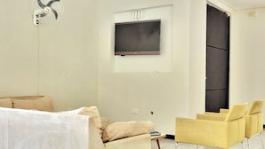 A modern room with a beige sofa, a yellow armchair, a flat-screen TV mounted on the wall, and a ceiling fan.
