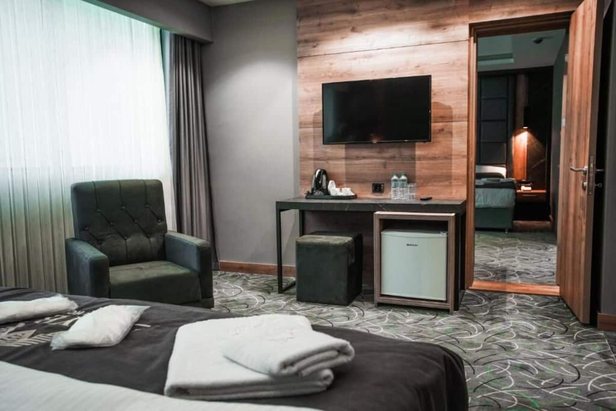 Habitación de hotel con cama, escritorio con televisor de pantalla plana, una silla, refrigerador pequeño y baño visible a través de una puerta abierta.