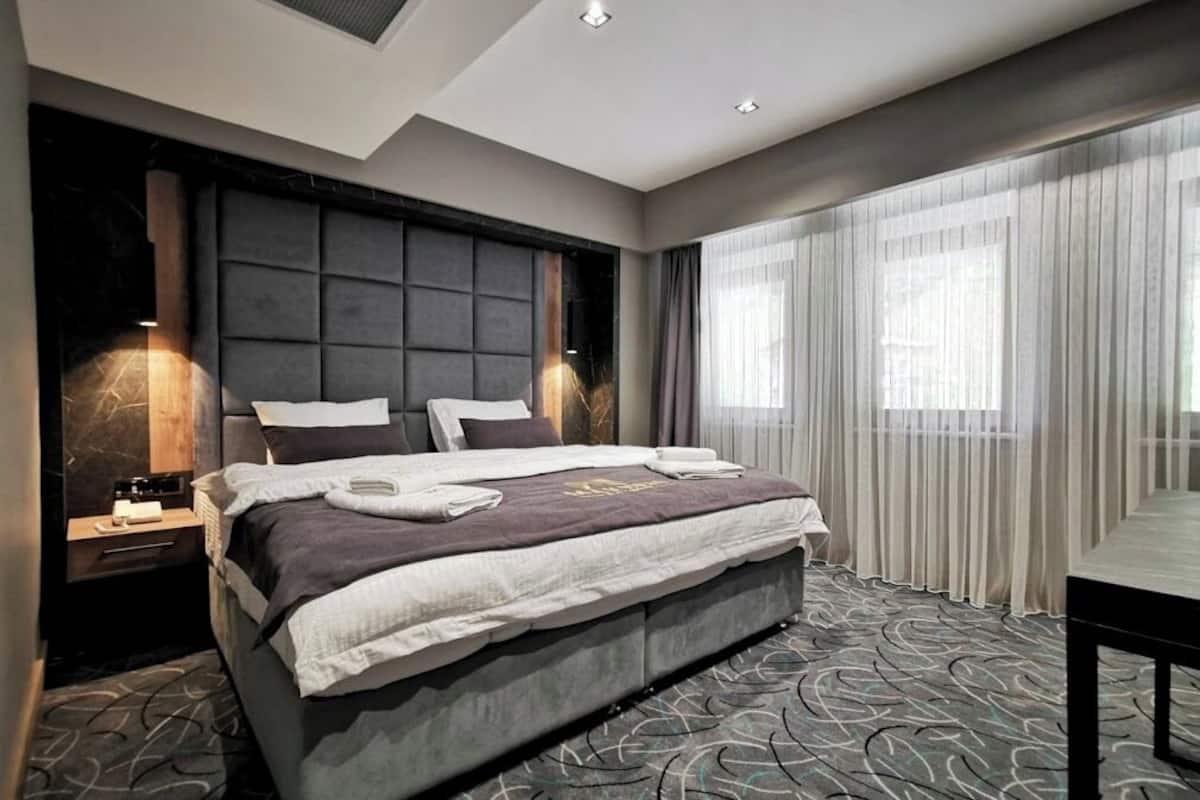 Una habitación de hotel moderna con una cama grande, una mesita de noche, un escritorio y vistas al exterior a través de cortinas translúcidas.