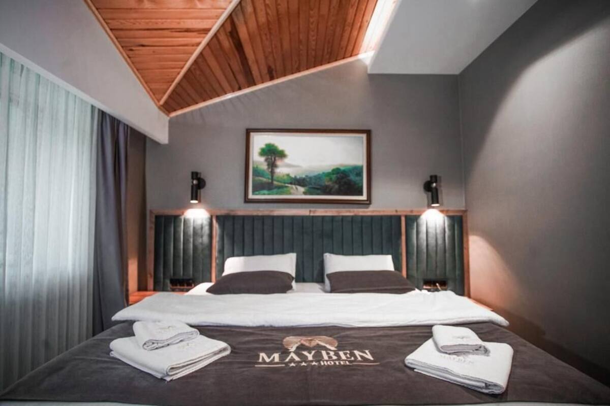 Habitación de hotel con una cama, dos mesitas de noche, cabecera, un cuadro y techo con vigas de madera.