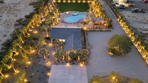 An aerial view of a resort with a swimming pool, a covered patio, and landscaped areas with palm trees and lights.