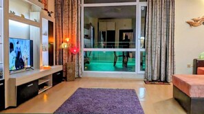 A living room with a television, a purple rug, and a view of a balcony with a pool. - Sunny Beach Serenity Apartment (Nessebar)