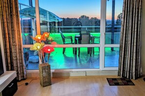 Una habitación con un ventanal grande, piso de madera, una pequeña mesa con sillas y un jarrón decorativo con flores.