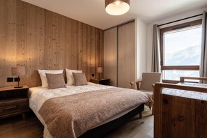 A bedroom with a wooden wall, a bed with a patterned blanket, bedside lamps, a chair, and a window with a view. - 50m from ski slopes, 1st floor, view resort, balcony, tv, 86m², Montalbert (La Plagne)