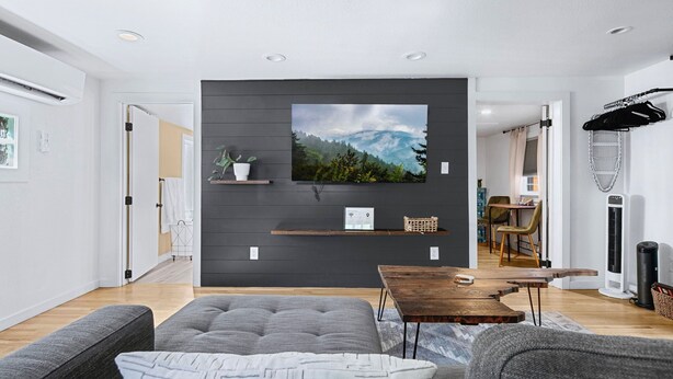 A modern living room with a flat-screen TV mounted on a dark wall, a wooden coffee table, grey sofas, and a dining area in the background. - Earthy Pearl - 1 Bedroom Suite @ the Boise Depot (Boise)