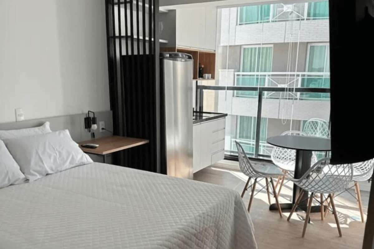 A modern hotel room with a bed, a small desk, a chair, and a balcony with a table and chairs.