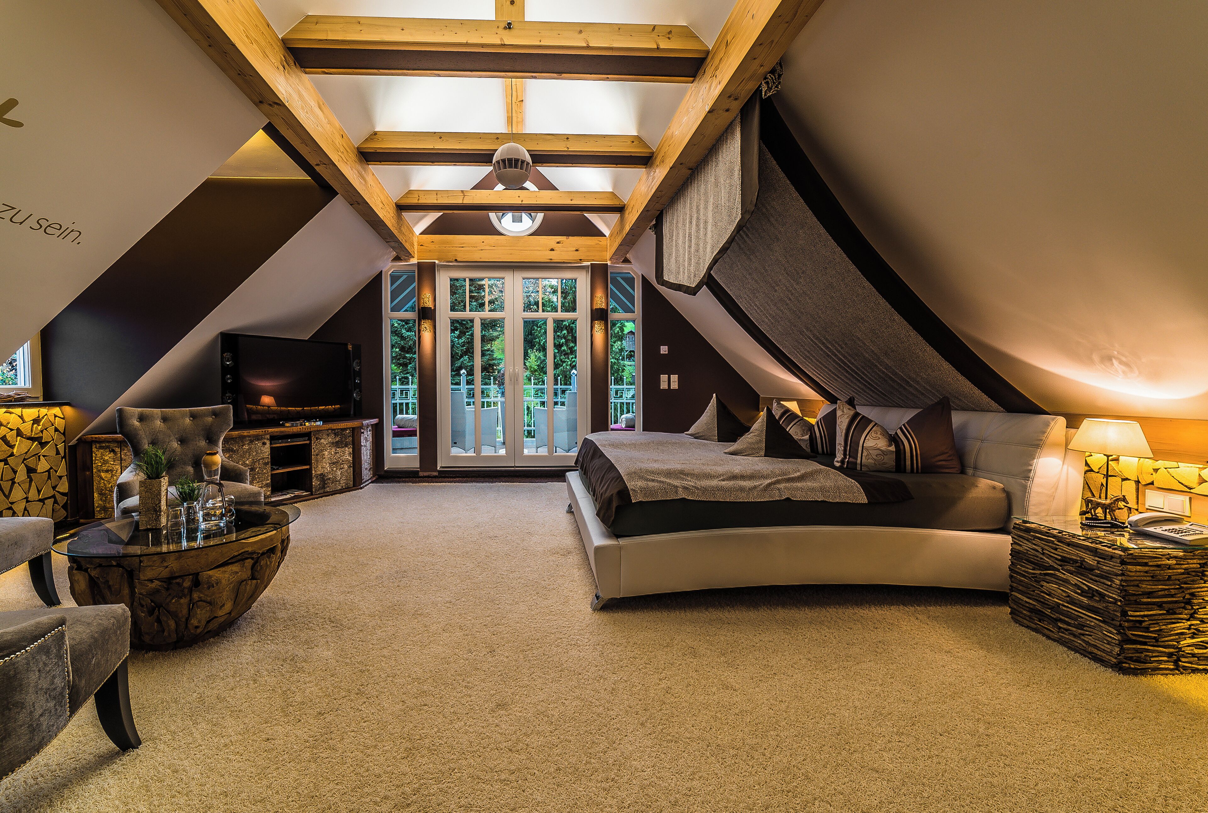 A cozy attic bedroom with a curved bed, a plush armchair, a glass coffee table, and a TV.