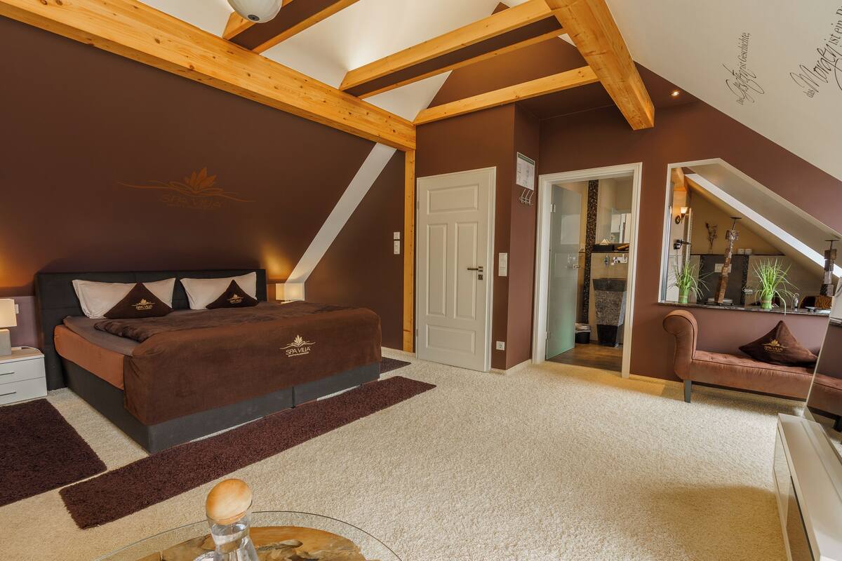 Une chambre dotée d’un grand lit, de poutres apparentes, d’une porte donnant sur une salle de bain et d’un petit coin salon avec un canapé.