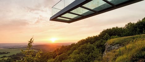 Un pont moderne en verre enjambant une colline boisée, au coucher du soleil.