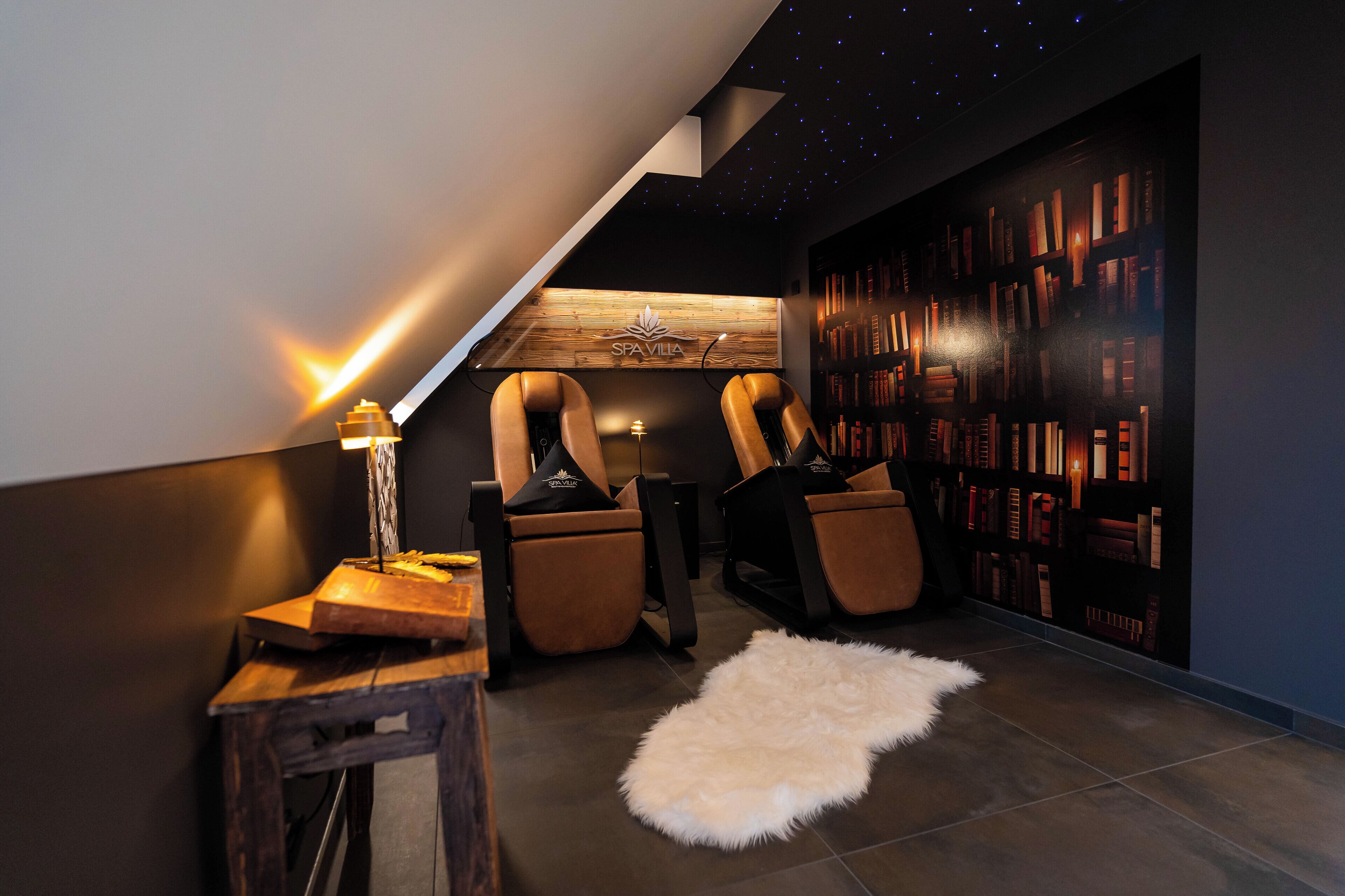 Un coin lecture moderne avec un plafond en pente, un mur en bois portant l’inscription « SPA VILLA », des fauteuils en cuir, une petite table avec une lampe et une bibliothèque.