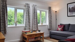 A cozy living room with a grey sofa, a wooden coffee table with board games, and a view of greenery through large windows with patterned curtains.