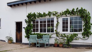 A white building with a black roof, two windows with white frames, and a door. There are two green chairs outside and several potted plants.