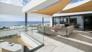 Uma área externa moderna para relaxar, com guarda-corpo de vidro, sofás bege e vista para o mar.