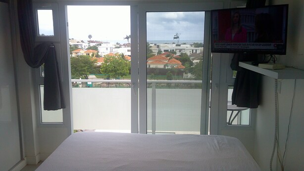 A hotel room with a bed, a TV mounted on the wall, and a balcony with a view of the city. - E&J Residences at SOV (Cabarete)