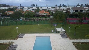 A swimming pool with lounge chairs, a fenced sports area, and residential buildings in the background. - E&J Residences at SOV (Cabarete)