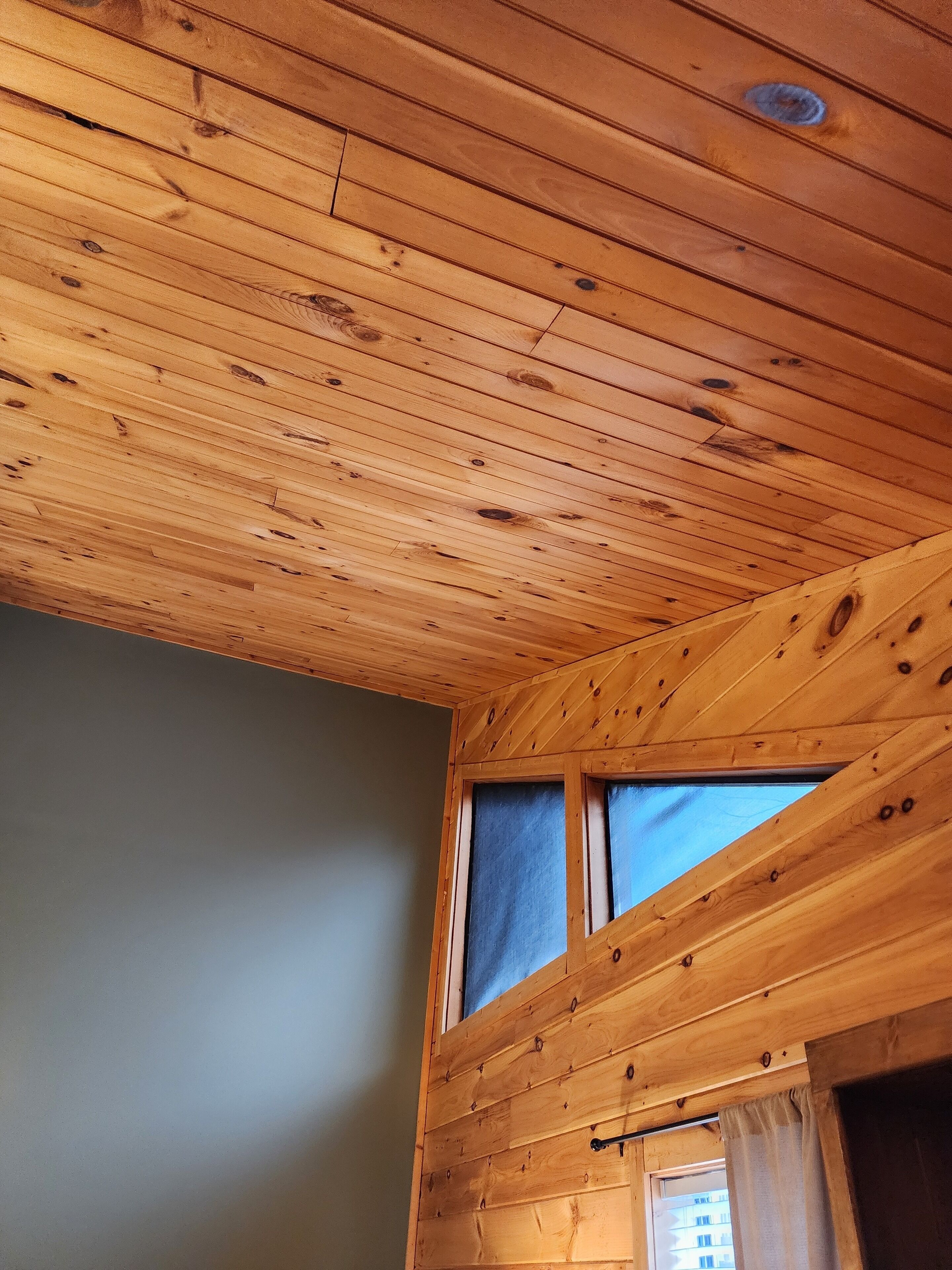 A room with wooden ceiling planks, a window with blue curtains, and a view of buildings outside.