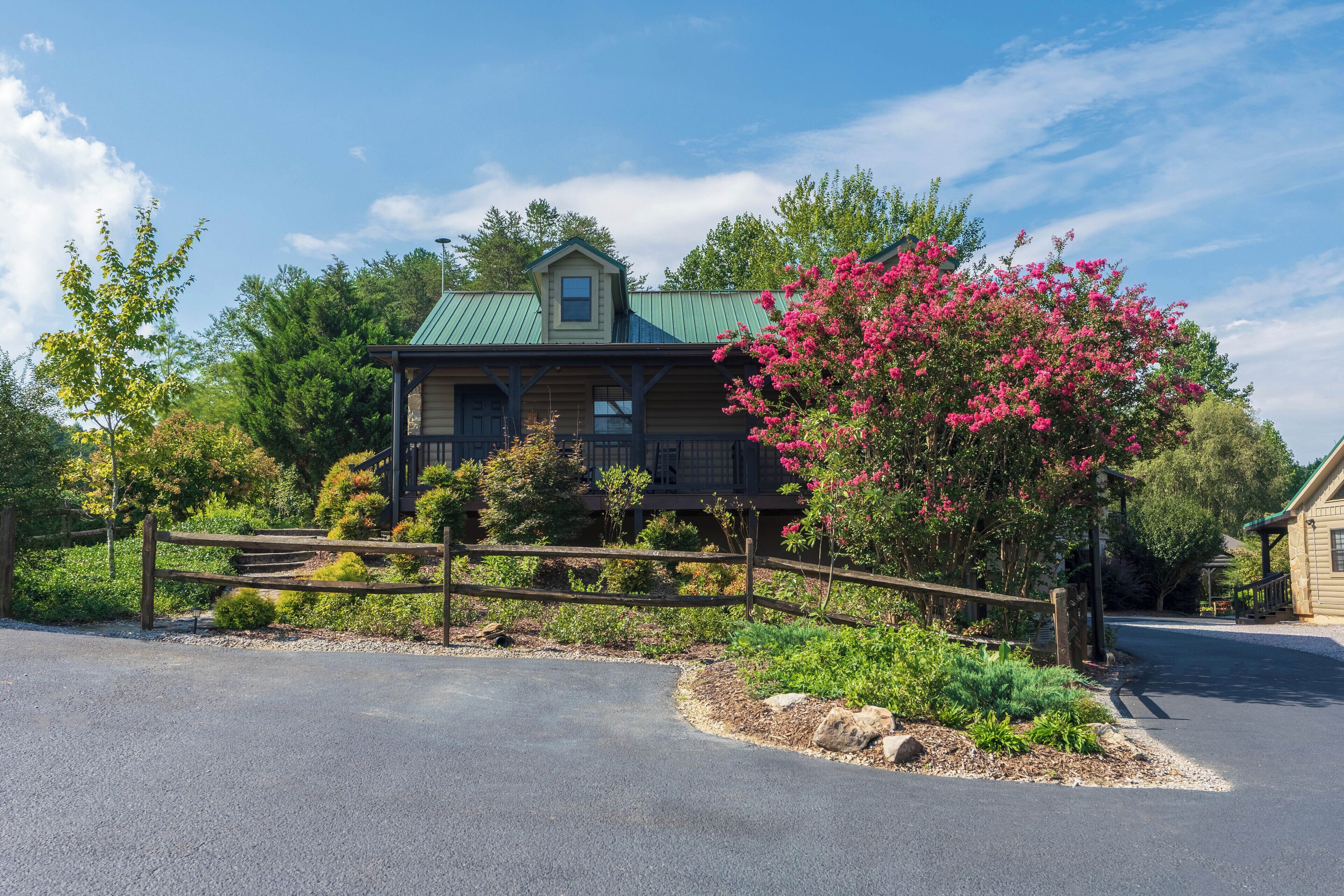 A wooden house with a green roof, surrounded by a wooden fence and a garden with pink flowers.