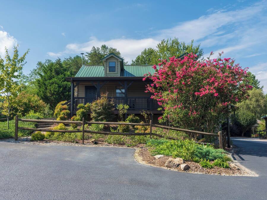 A wooden house with a green roof, surrounded by a wooden fence and a garden with pink flowers.