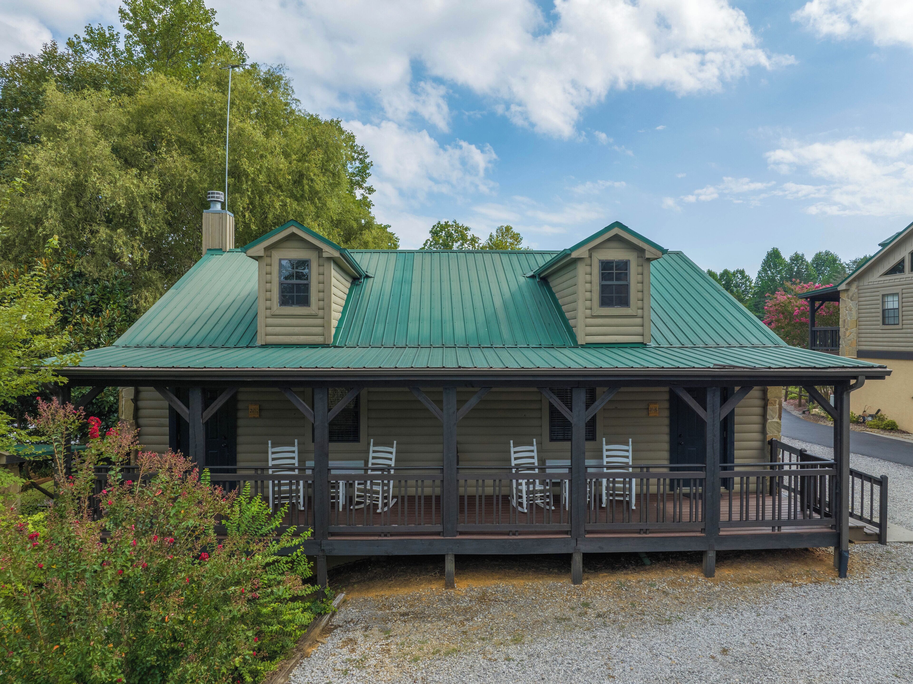 A two-story log cabin with a green metal roof, a porch with white chairs, and a stone chimney.