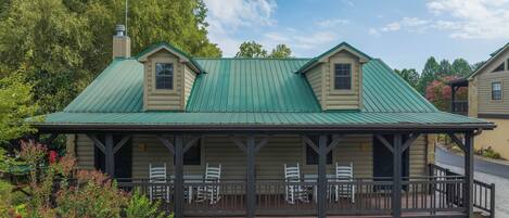A two-story log cabin with a green metal roof, a porch with white chairs, and a stone chimney.