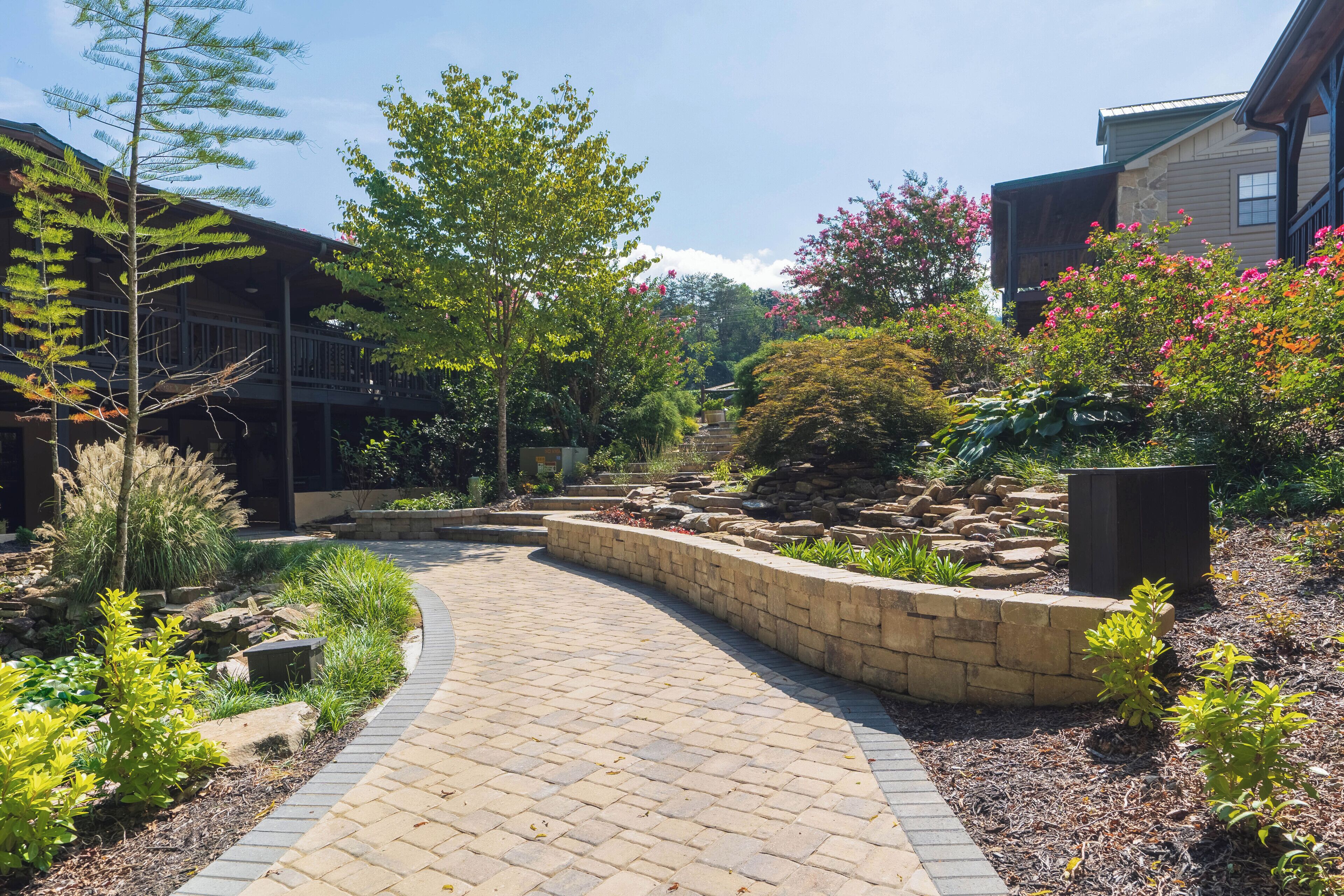 A paved pathway leading to a building with a stone retaining wall and various plants.
