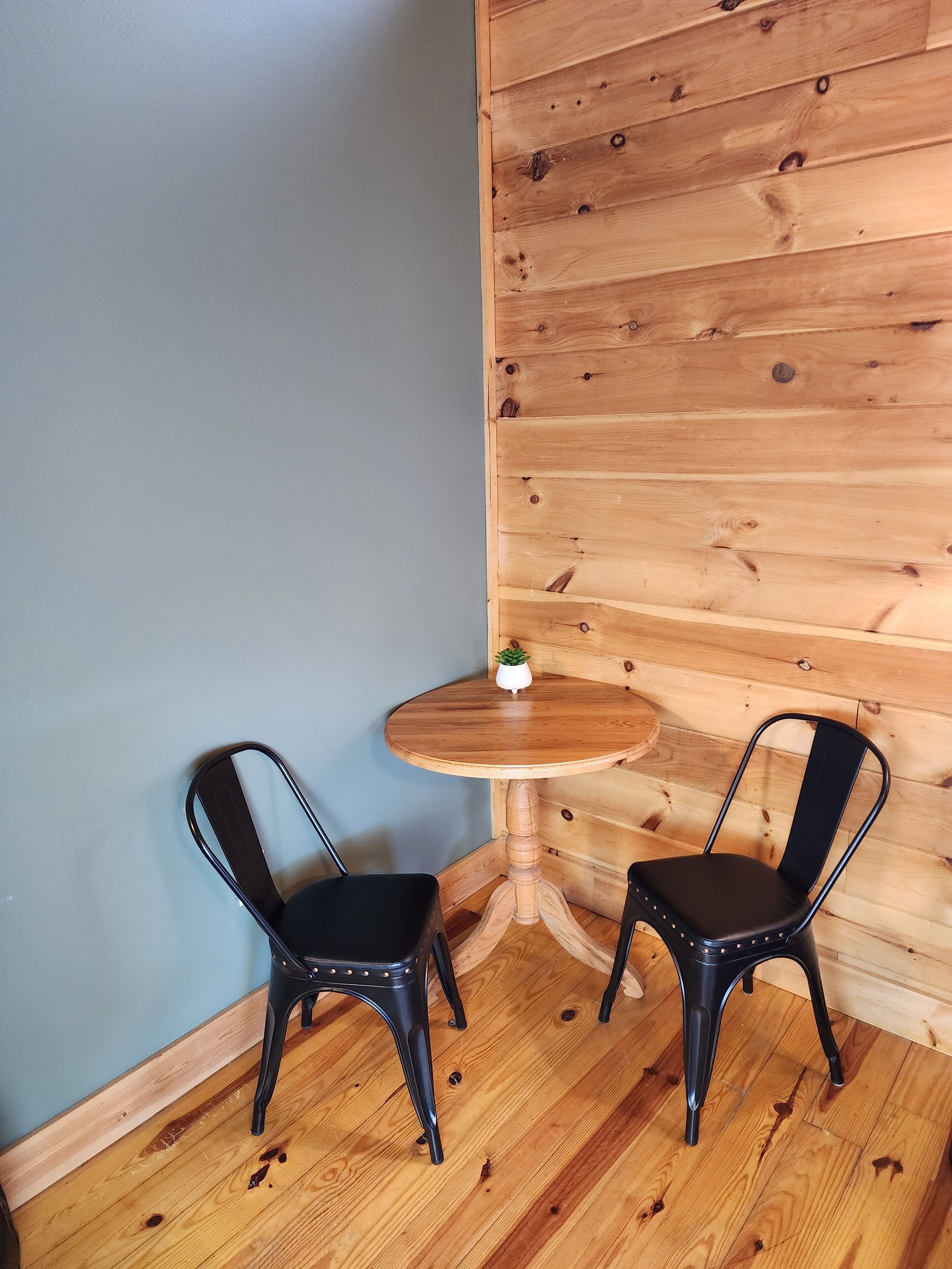 A corner room with a wooden floor, two black chairs, a small round wooden table, and a potted plant.