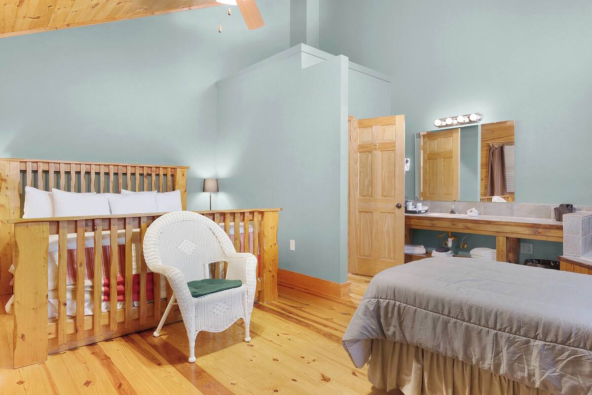 A room with a crib, a white wicker chair, a bed, a wooden dresser, and a mirror with lights.