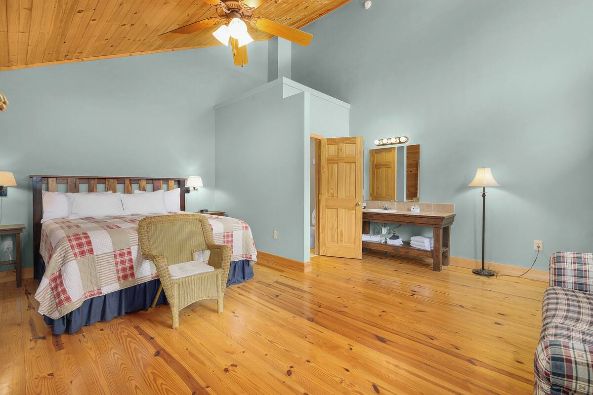A bedroom with a wooden ceiling, a bed with a checkered bedspread, a chair, a bedside table, a nightstand, a mirror, and a floor lamp.