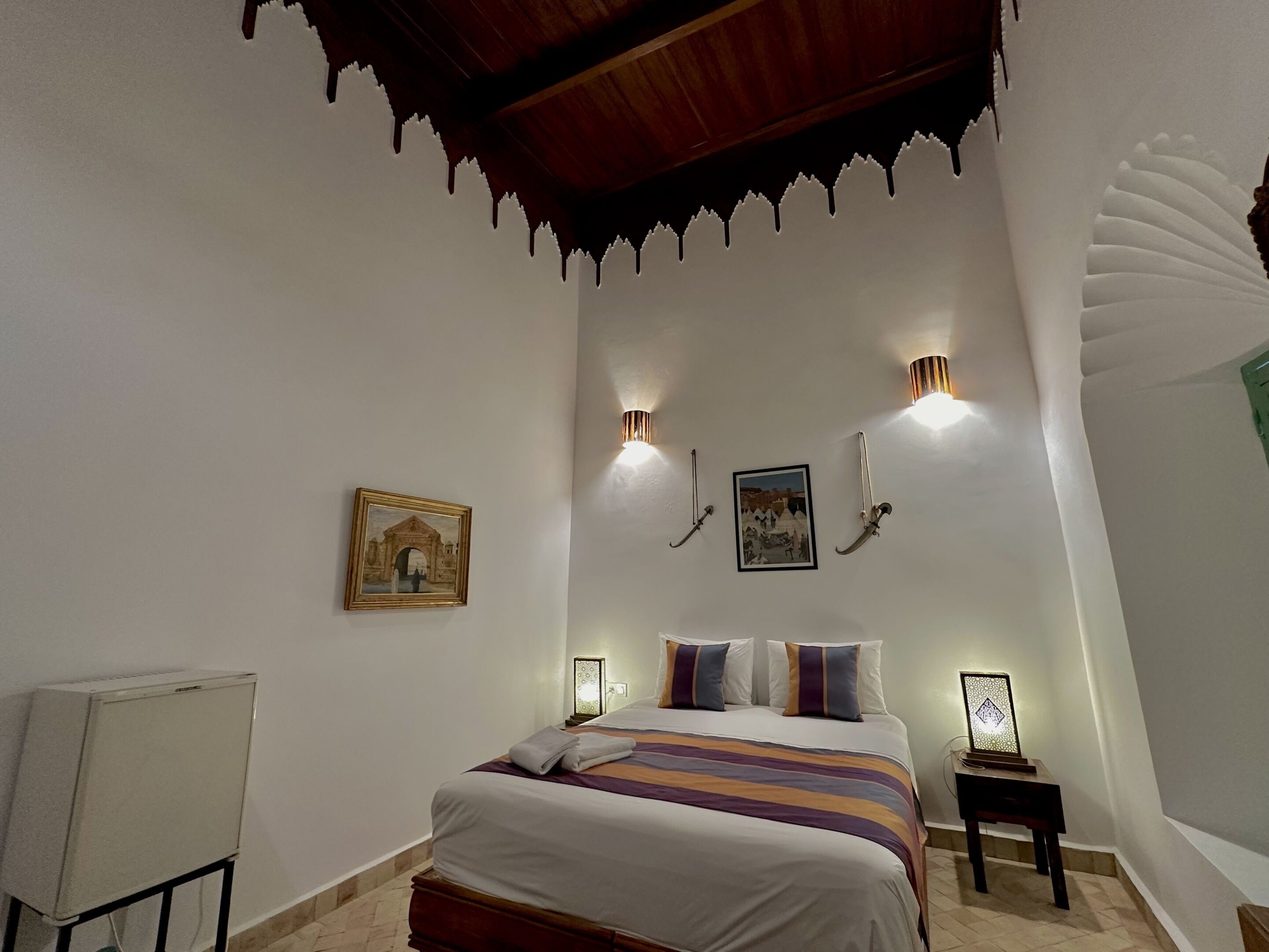 A bedroom with a bed, two wall-mounted lights, a framed picture, and a small table with a lamp.