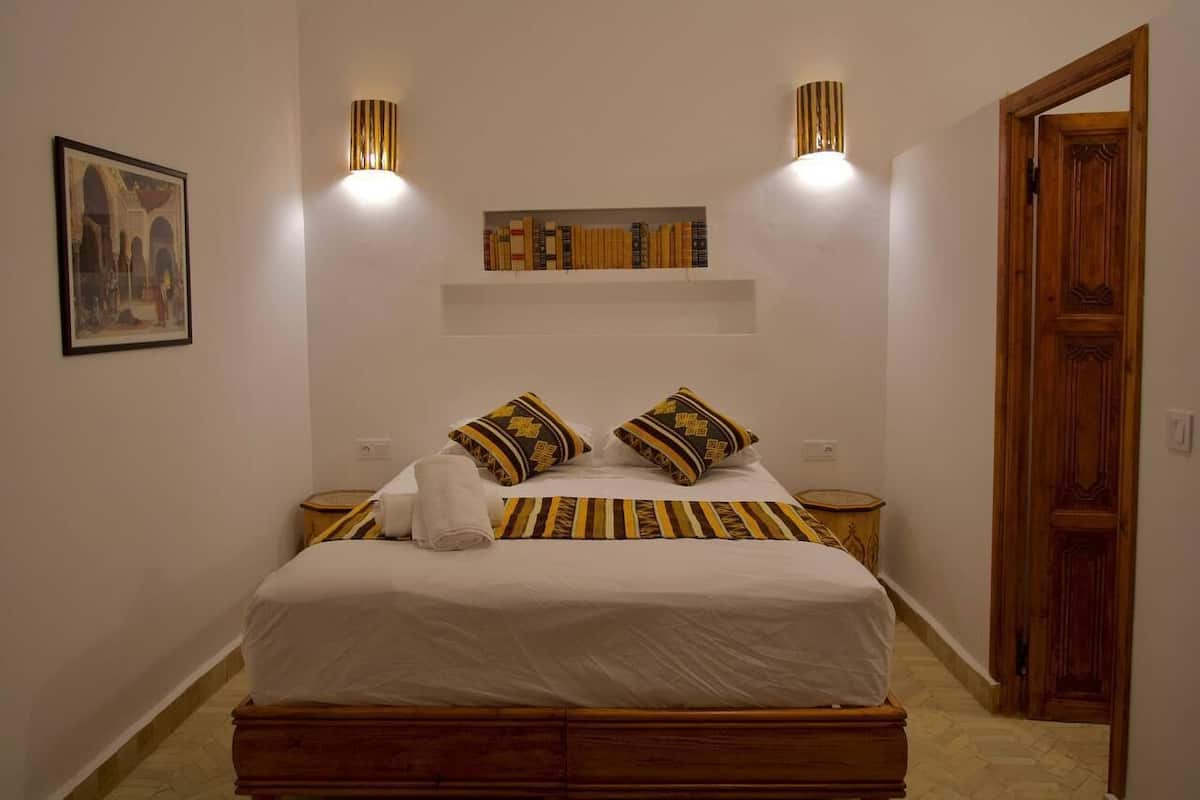 A bedroom with a bed, two bedside tables, a wall-mounted shelf with books, a framed picture, and a wooden door.