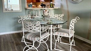 Un comedor con una mesa de vidrio y sillas de metal blanco, un reloj decorativo en la pared y un espejo que refleja un balcón.