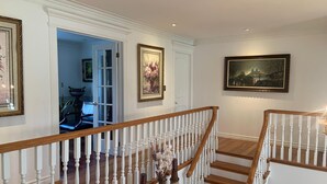 Un escalier avec des balustrades blanches et des rampes en bois, menant à un palier orné d’un vase de fleurs et d’un tableau encadré.