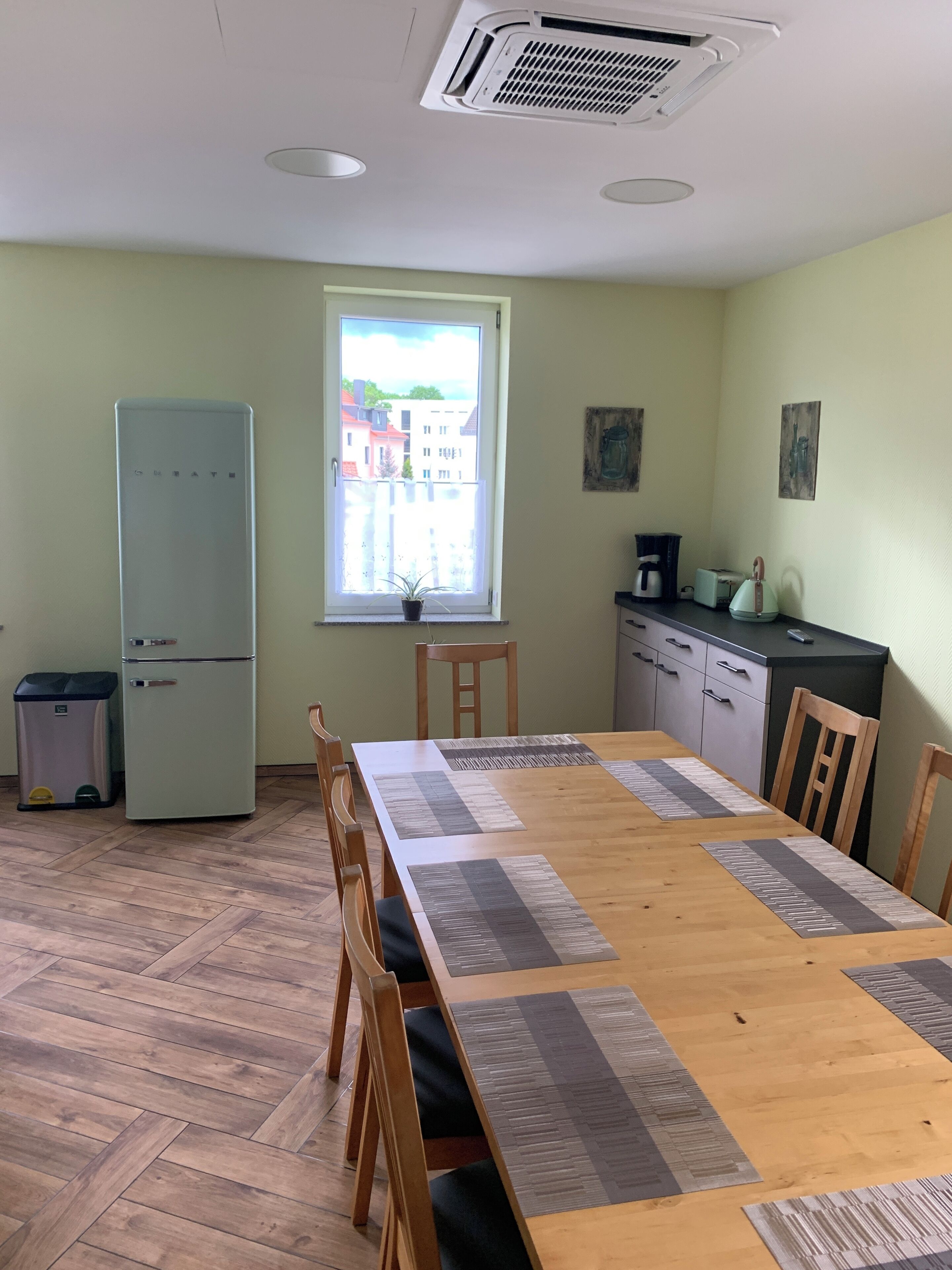 Un espace repas comprenant une table en bois, des chaises, un réfrigérateur, une poubelle, une fenêtre donnant sur des immeubles, ainsi qu’un coin cuisine avec des placards et des appareils électroménagers.