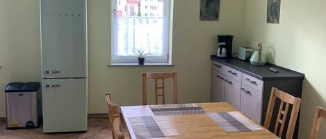 Un espace repas comprenant une table en bois, des chaises, un réfrigérateur, une poubelle, une fenêtre donnant sur des immeubles, ainsi qu’un coin cuisine avec des placards et des appareils électroménagers.