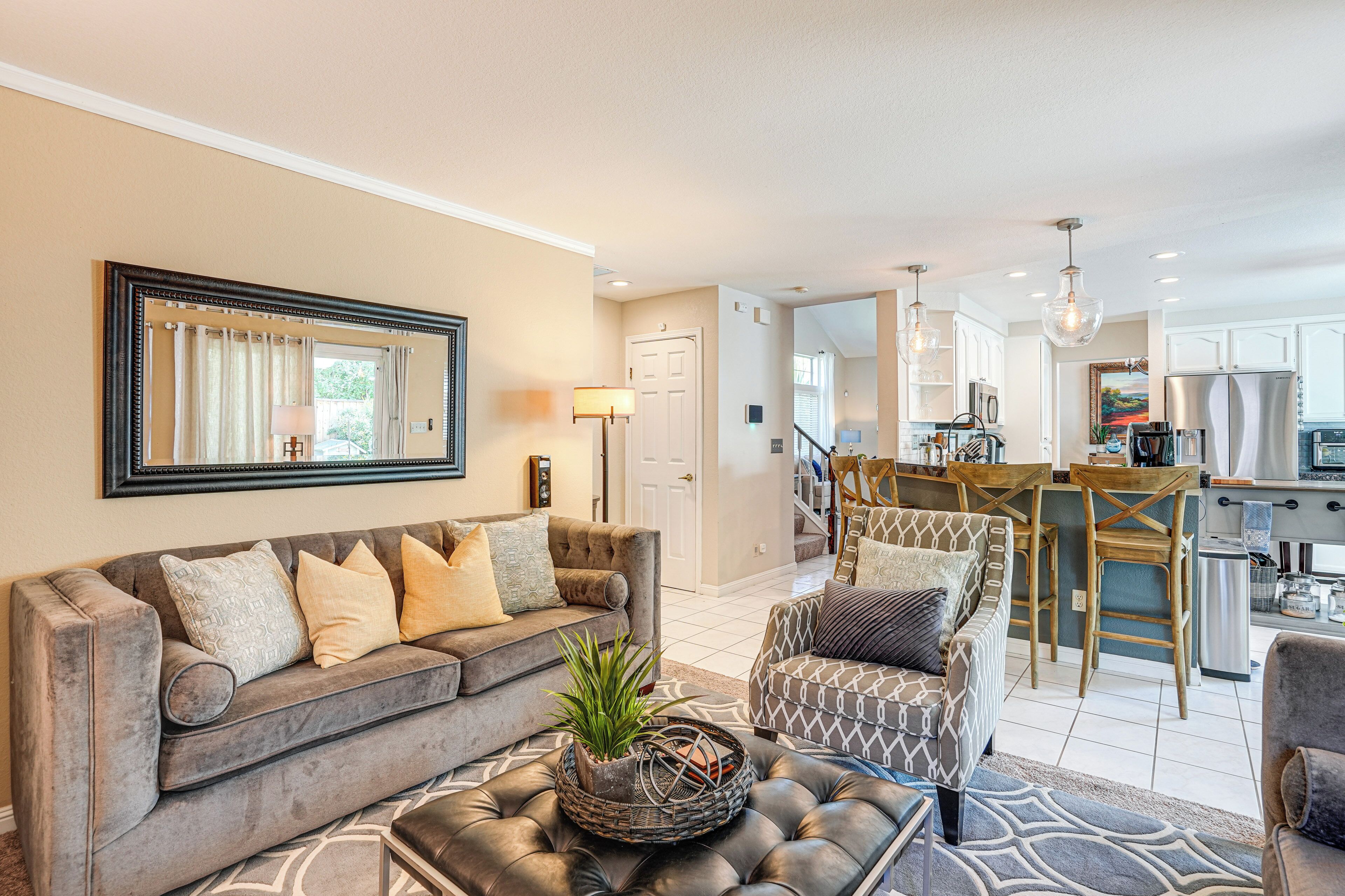 Ein geräumiges Wohnzimmer mit grauem Sofa, gemustertem Sessel und Couchtisch mit Pflanze. Im Hintergrund ist eine Küche mit modernen Geräten und Barhocker zu sehen.