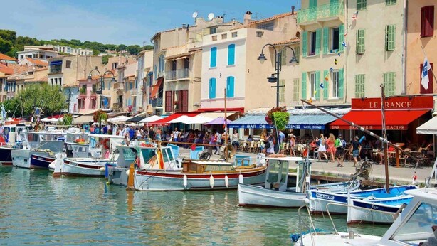 Ein Yachthafen mit vertäuten Booten, eine belebte Uferpromenade mit Menschen und Gebäude mit Geschäften und Restaurants.