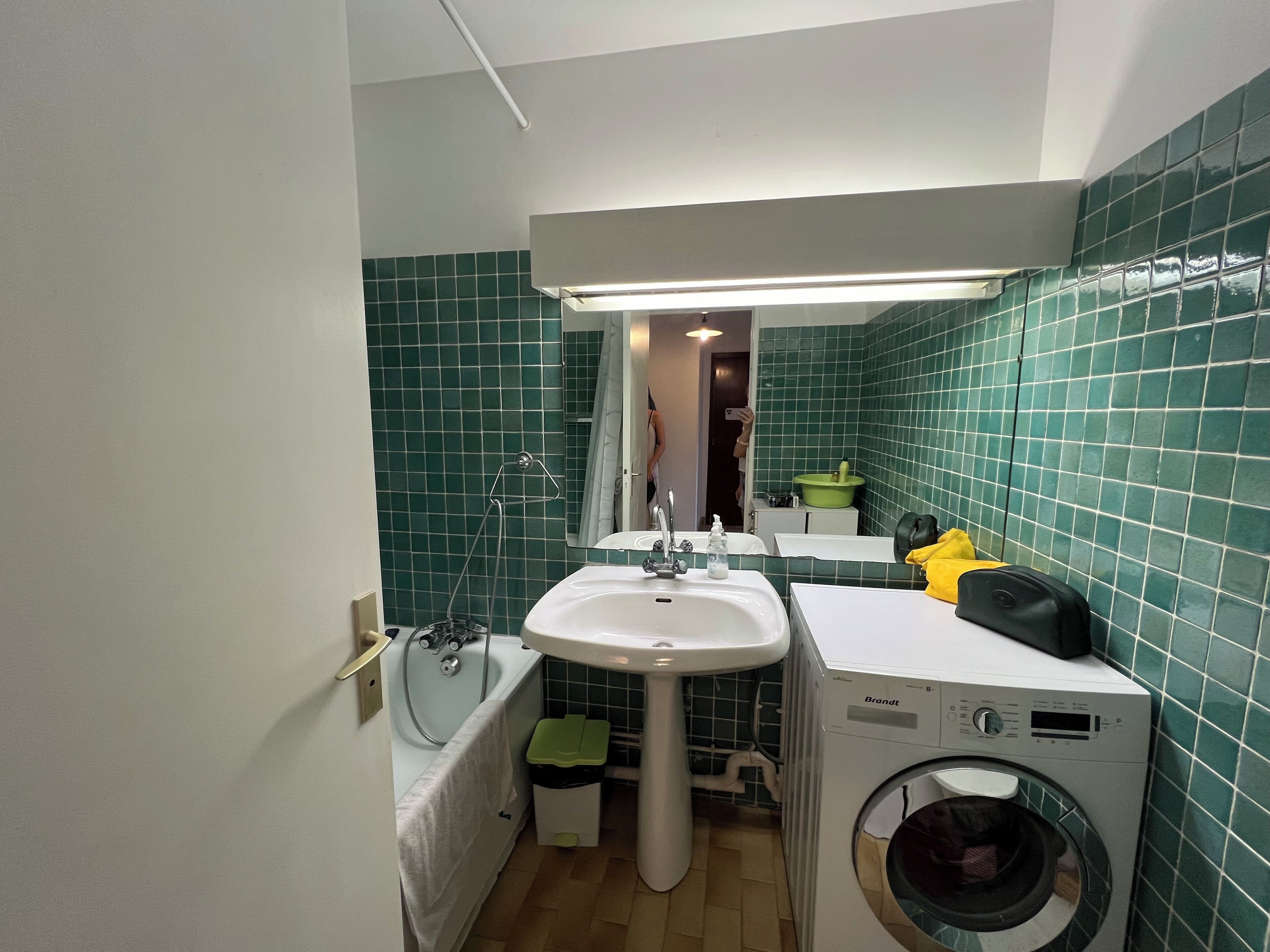 A compact bathroom with a sink, mirror, green tiled walls, a bathtub, and a front-loading washing machine.