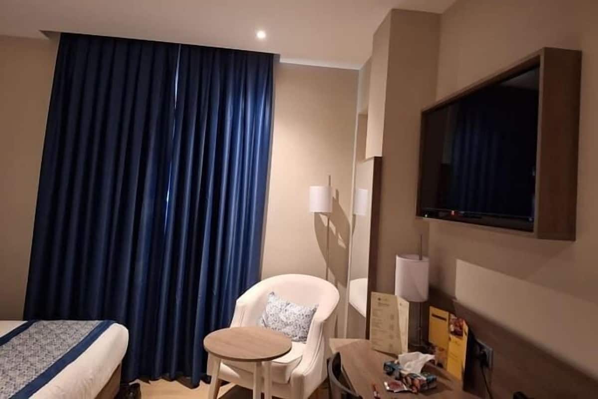 Hotellihuone, jossa on sänky, pieni pöytä, tuoli, televisio ja ikkuna, jossa on verhot.