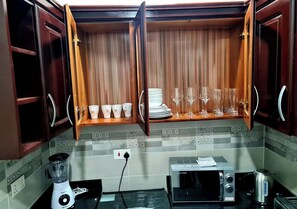 A kitchen with wooden cabinets, a microwave, a toaster, a blender, and a shelf with cups and glasses. - Charming Quiet & Cozy Close to Highway (North Legon)
