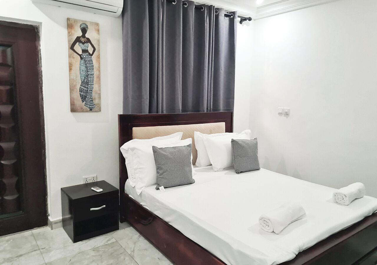 A neatly made bed with white linens and pillows, a small bedside table with a remote control, and a painting on the wall.