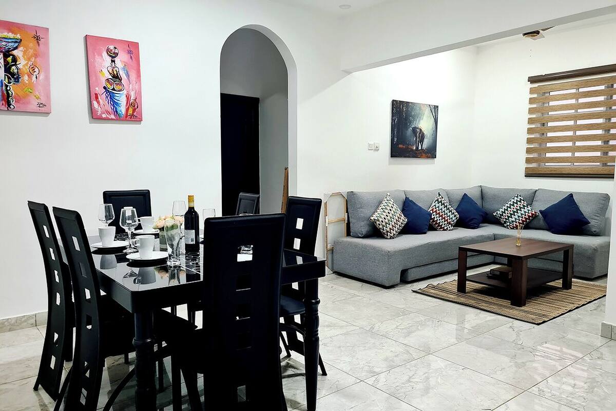 Un comedor con una mesa dispuesta para cuatro personas, un sofá gris con cojines estampados y un cuadro en la pared.