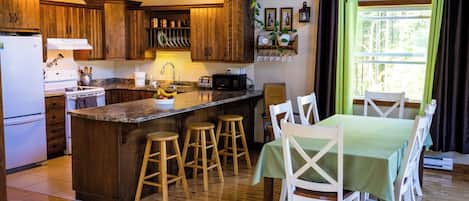 A kitchen with wooden cabinets, a central island with stools, a dining table with chairs, and a window with curtains.
