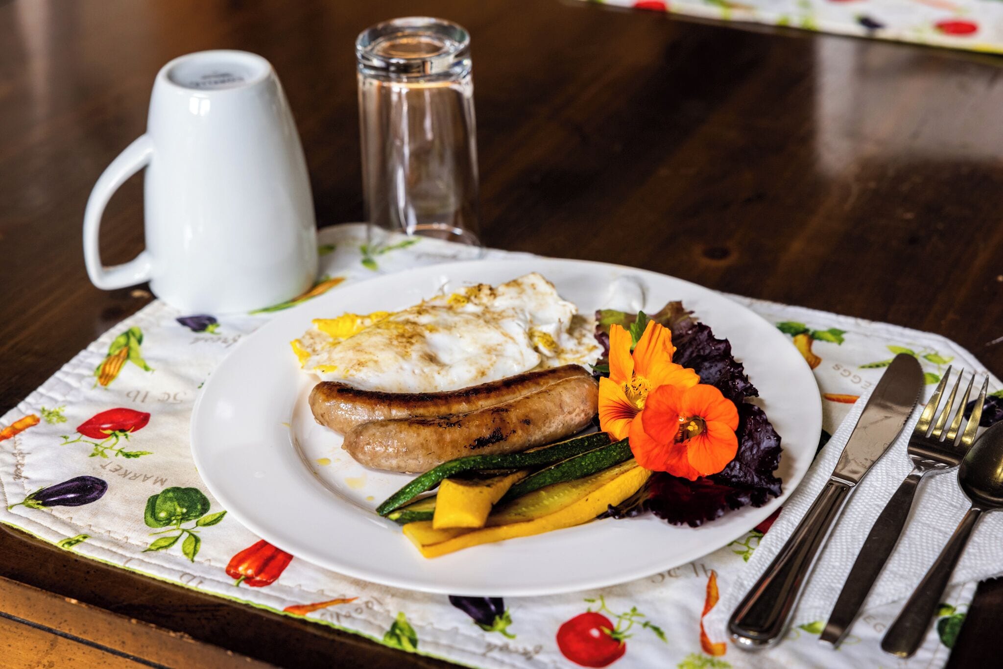 Une assiette remplie de nourriture, une tasse blanche et des couverts sont posés sur une table recouverte d’une nappe à motifs floraux.