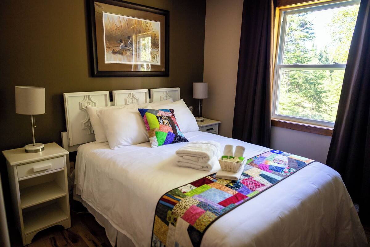 A neatly made bed with a colorful quilt, a bedside table with a lamp, and a window with curtains.