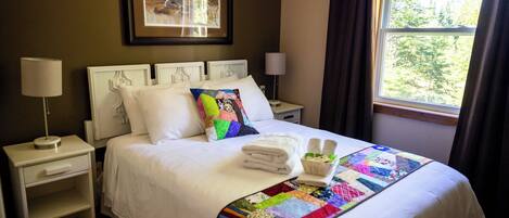 A neatly made bed with a colorful quilt, a bedside table with a lamp, and a window with curtains.