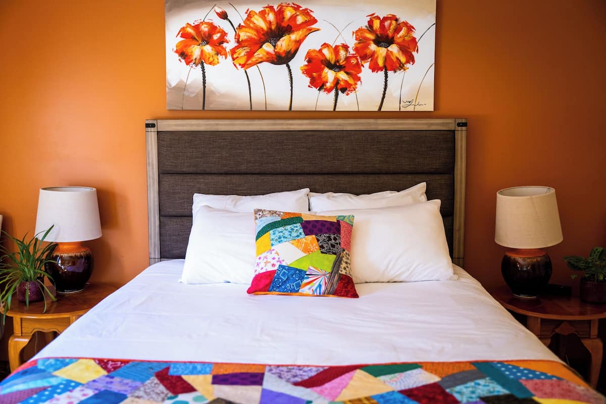 A neatly made bed with a colorful quilt, a large floral painting above it, and two bedside lamps on wooden nightstands.