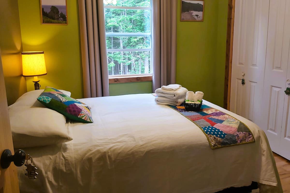 A neatly made bed with a quilt, pillows, and a folded towel set on a bed with a view of trees through a window.