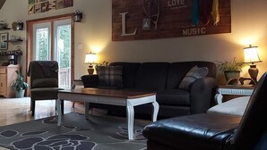 Ein geräumiges Wohnzimmer mit hoher Decke, einem großen L-förmigen Sofa, einem Couchtisch und einem gemusterten Teppich. Zwei Stehlampen, ein Holz-Wandbild und eine Tür zum Garten sind ebenfalls vorhanden.